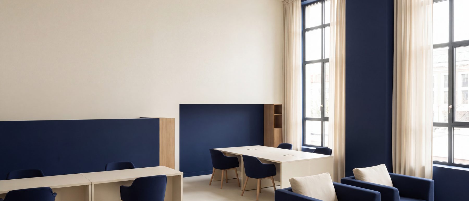 An office with cream walls and navy panels, workstations and a lounge area under tall windows